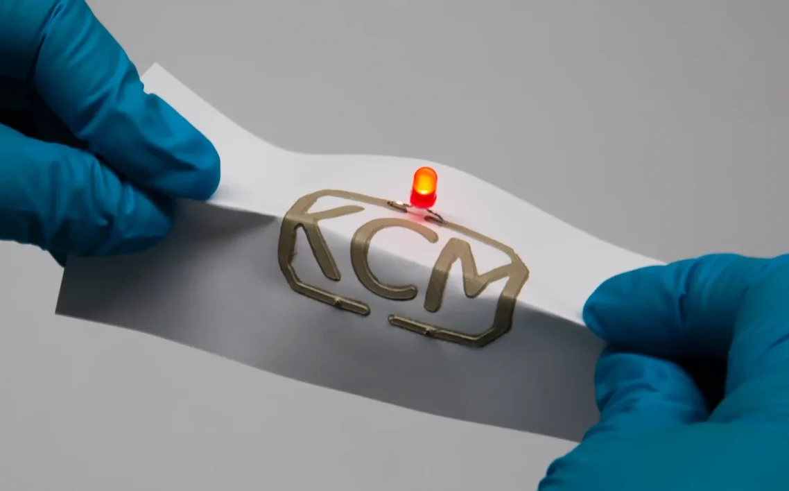 A person wearing blue gloves bends a sheet bearing the KCM logo and a red, glowing LED.
